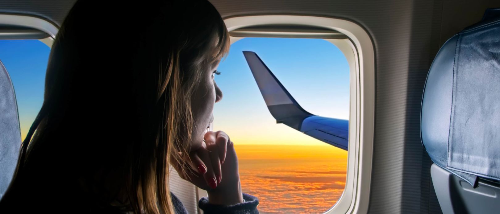 A young beautiful girl looks out the window of the plane to the fantastic clouds at sunset. The passenger looks out the window of the plane to the wing. Travel, vacations, holiday and tourism concept